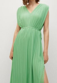 Vestido verde plisado con escote en V, mangas sueltas y cintura ceñida con un cinturón. La tela tiene una textura suave y un diseño fluido.