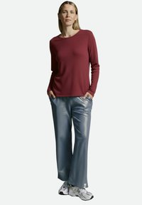 Dunkelrotes Langarmoberteil mit strukturierter Oberfläche, kombiniert mit glänzenden, locker sitzenden grauen Hosen mit dezentem Muster und Taschen. Weiße Sneakers.