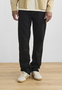 Pantalones de mezclilla negros de corte recto, combinados con una camisa de pana color beige claro y zapatillas blancas con rayas marrones.
