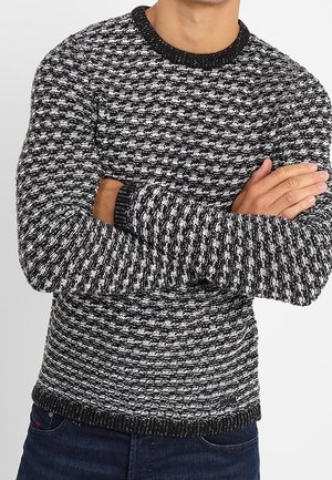 Man draagt een zwarte, grijze en witte gebreide trui met lange mouwen, staat met gekruiste armen tegen een effen achtergrond.