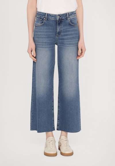 Hellblaue Jeans mit weitem Bein und hoher Taille. Besitzt fünf Taschen, Gürtelschlaufen und einen ausgewaschenen Look; kombiniert mit beigen Turnschuhen.