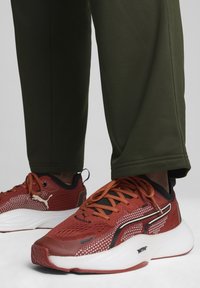 Baskets de sport rouges avec détails en maille, accents noirs et semelle blanche. Présentées avec un pantalon vert olive, mettant en valeur le design de la chaussure.