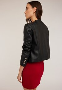 Czarna skórzana kurtka o skróconym fasonie, z złotymi guzikami na rękawach i gładką fakturą, zestawiona z czerwoną spódnicą ołówkową.