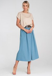 Jasno-beżowa bluzka z krótkim rękawem w połączeniu z niebieską midi spódnicą z wysokim stanem i fałdami oraz szerokim czarnym paskiem; modelka nosi beżowe sandały na kostkę z paskiem.
