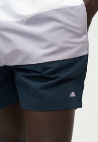 Σκούρα μπλε αθλητικά σορτς που φοριούνται από ένα άτομο με ανοιχτόχρωμο πουκάμισο, με ένα μικρό ανοιχτό ροζ ημικυκλικό λογότυπο κοντά στο τελείωμα.