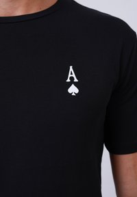 T-shirt en coton noir avec un 'A' imprimé blanc et un symbole de trèfle sur la zone gauche de la poitrine ; manches courtes, texture lisse.