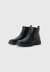 Botas de tobillo de cuero negro con un acabado liso, cremallera lateral y suela de goma resistente con un patrón de relieve. Dos tiradores en la parte posterior.