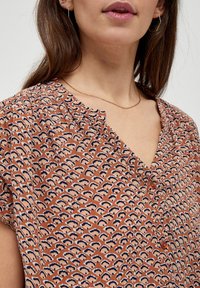Donna che indossa una blusa a motivi color ruggine con bottoni e una collana d'oro, in cui si possono vedere i dettagli della parte inferiore del volto e del collo.