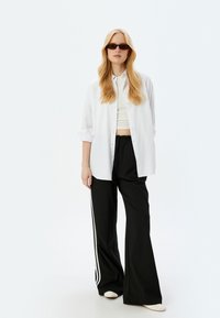 Camisa blanca de botones, top corto blanco, pantalones negros de pierna ancha con rayas laterales blancas y gafas de sol. Texturas suaves y diseño casual.