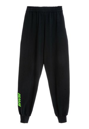 Joven mujer de pie mirando hacia adelante, vistiendo una camiseta negra de gran tamaño y pantalones negros holgados con detalles verdes sobre un fondo blanco liso.