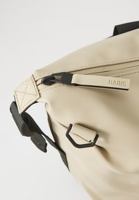 Beige waterproof backpack with a black zipper and adjustable strap, featuring a textured finish and logo detail on the zip pull.