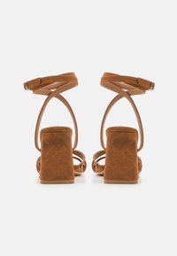 Sandales à talons hauts en daim marron avec un large talon bloc, dos ouvert et brides entrecroisées à la cheville. Texture lisse et design minimaliste.