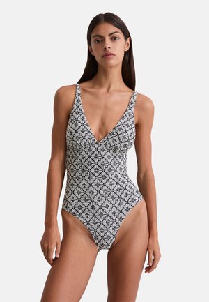Femme portant un maillot de bain une pièce à motifs floraux noir et blanc, avec un décolleté en V profond et des fines bretelles.