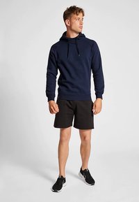 Marineblå hættetrøje med tekstureret finish, kombineret med sorte shorts. Modellen bærer sorte sneakers og står foran en neutral baggrund.