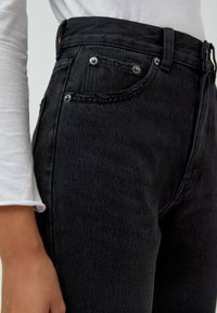 Jeans de mezclilla negros de tiro alto con herrajes plateados, que presentan un bolsillo frontal y una tela texturizada. El diseño incluye detalles de costura sutiles.