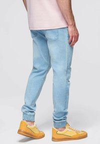 Světle modré denimové joggery s manžetami u kotníků, designem s pěti kapsami a uvolněným střihem, zkombinované se žlutými a bílými teniskami.