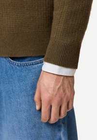 Männliche Hand ruht neben blauen Jeans, trägt einen olivgrünen Strickpullover über einem weißen Hemd, dessen Manschette sichtbar ist.