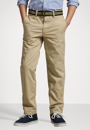 Enfant portant un pantalon beige, des baskets bleu marine avec des lacets blancs, une ceinture rayée et une chemise à manches longues rayée bleu et blanc avec les manches retroussées.