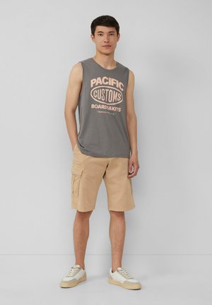 Graues ärmelloses Shirt mit dem Schriftzug "PACIFIC CUSTOMS" in Pfirsich, kombiniert mit hellbeigen Cargo-Shorts und weißen Sneakers. Einfaches, lässiges Outfit.