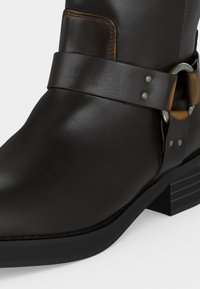 Μαύρο δερμάτινο μποτάκι αστραγάλου με λουράκι και ασημένια αγκράφα στη στιβαρή φτέρνα, σε λευκό φόντο.