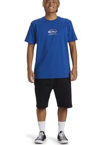 Camiseta de algodón azul con cuello redondo, mangas cortas y un pequeño gráfico en el pecho. Combinada con pantalones cortos negros y zapatillas deportivas.