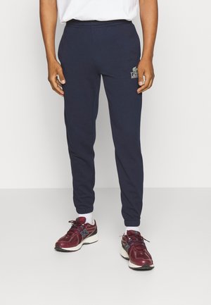 Mand iført marineblå Lacoste joggingbukser, hvide sokker, burgundy New Balance-sneakers og en hvid skjorte, stående på et ensfarvet gulv.