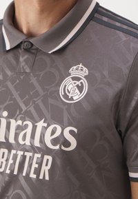 Zbliżenie szarego stroju piłkarskiego Real Madryt z białym haftowanym logo oraz napisem sponsora Emirates BETTER na teksturowanej tkaninie.