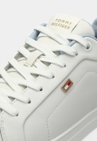Sneaker in pelle bianca con lacci bianchi, rivestimento azzurro chiaro, logo Tommy Hilfiger sulla linguetta e piccolo emblema rosso-bianco-blu sul lato.