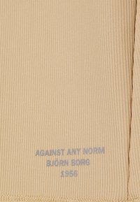 Béžová ribkovaná tkanina s hladkým povrchem, na které je blízko lemu modrý potištěný text: "PROTI JAKÉMKOLI NORMÁM BJÖRN BORG 1958".