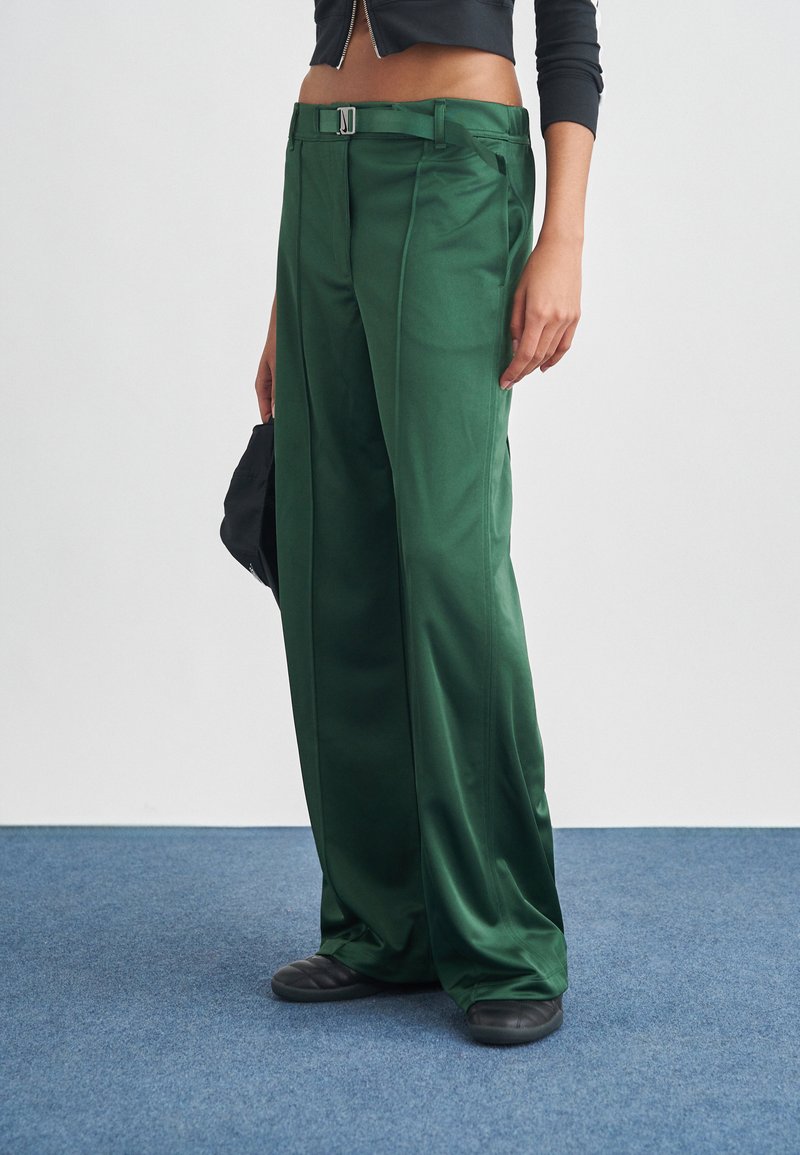 Πράσινο σατέν παντελόνι με φαρδύ πόδι, με πιέτες μπροστά, ασημένια αγκράφα ζώνης και λεία υφή, συνδυασμένο με μαύρα παπούτσια τύπου slip-on.