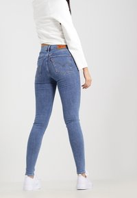 Vaqueros de mezclilla azul ajustados de cintura alta, con bolsillos traseros y una etiqueta de cuero de marca. Combinados con zapatillas blancas.