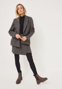 Blazer grigio in misto lana con una parte frontale a doppio strato, abbinato a un top nero aderente, una gonna scura e collant neri. Stivali alla caviglia marroni con lati elastici.