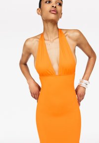 Robe orange à dos nu en tissu extensible, avec un décolleté en V profond et une silhouette ajustée. Les accessoires comprennent des bracelets transparents.
