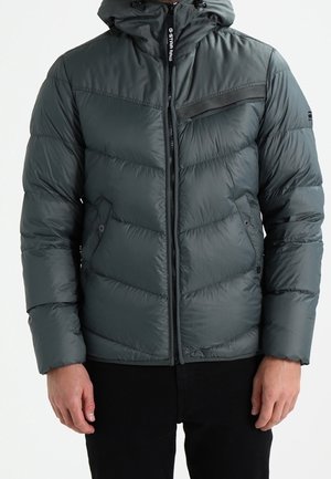 Mann trägt dunkelgraue gesteppte Winterjacke mit Kapuze, Frontreißverschluss, Reißverschlusstasche auf der Brust und Seitentaschen.