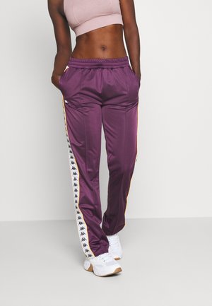 Persona con pantalones deportivos morados con franjas blancas de logotipo, top corto rosa y zapatillas blancas, de pie contra un fondo claro sencillo.