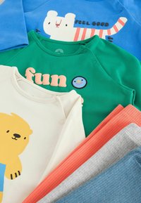 Cinq vêtements pour enfants : un haut bleu avec un graphique d'ours, un haut vert avec un texte et un smiley, un haut crème avec un imprimé d'ours, et deux paires de pantalons côtelés en orange et gris.