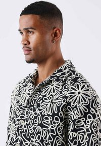 Camisa negra con patrón y diseños abstractos de flores en blanco, tela texturizada, cierre de botones y cuello de solapa.