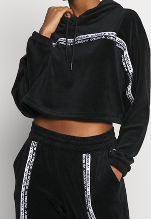 Persoon draagt een zwarte cropped hoodie en bijpassende broek met witte Adidas-logo strepen, hand raakt het koordje van de hoodie aan.