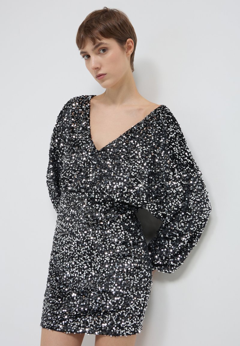 Abito mini nero in paillettes con scollatura a V profonda, maniche lunghe a sbuffo e superficie testurizzata. L'abito ha una silhouette aderente e dettagli luccicanti.