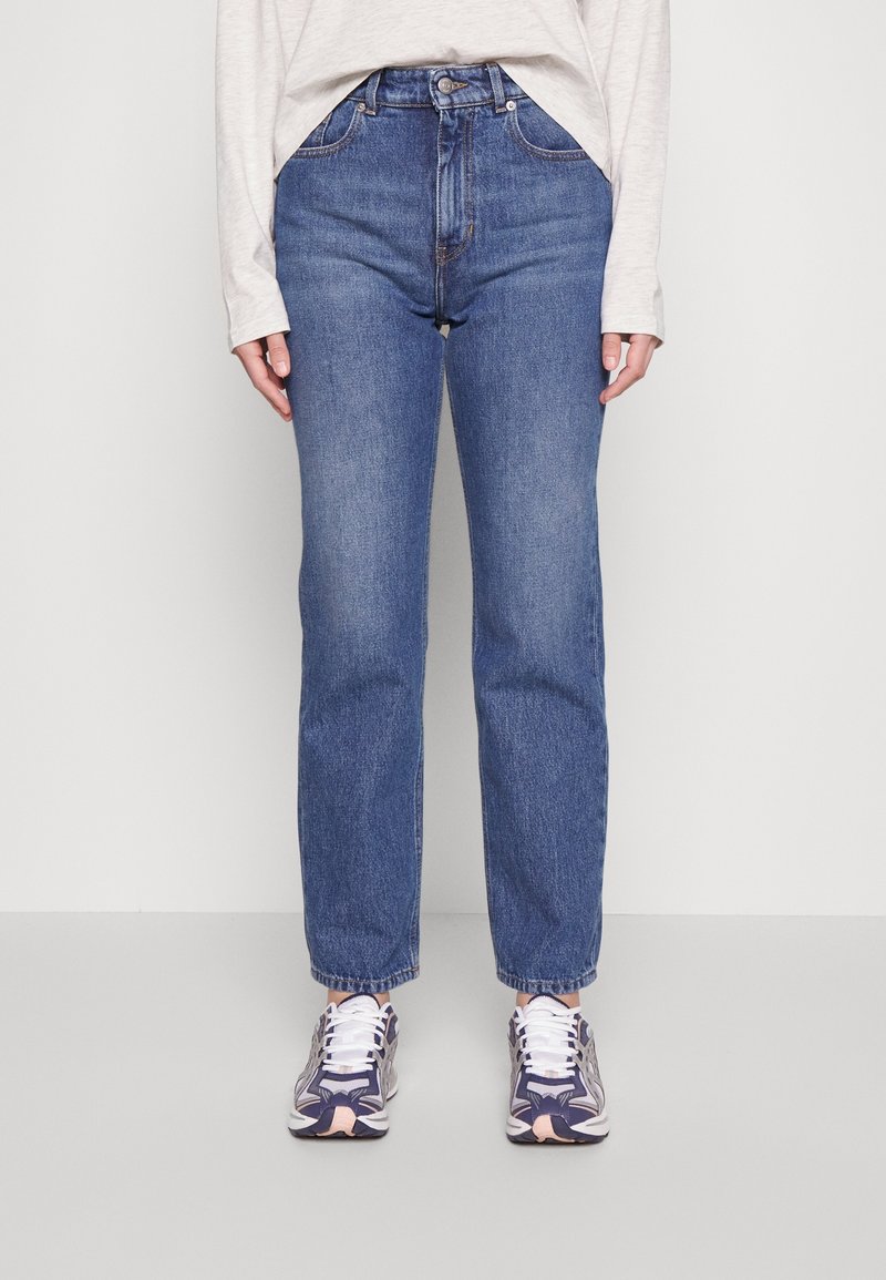 Personne portant un jean taille haute bleu, un t-shirt manches longues gris clair, et des baskets violettes et blanches, debout devant un fond blanc uni.