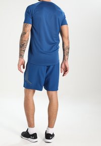 Krátký rukáv modré sportovní tričko s odpovídajícími šortkami, které mají kontrastní šedé akcenty a hladkou texturu. Černé sportovní tenisky s bílou podrážkou.