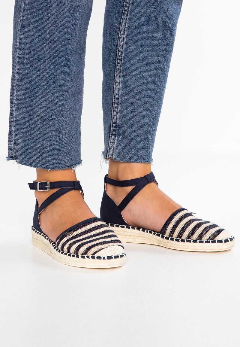 Pieds portant des espadrilles rayées bleues et blanches avec des brides à la cheville et un jean bleu court sur fond blanc.