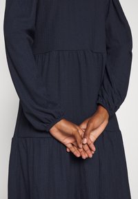 Mains d'une personne à la peau foncée croisées dans le dos, portant une robe longue à manches longues et à texture sombre.