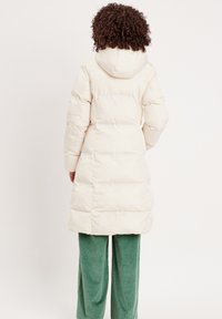 Manteau en duvet crème avec une capuche, présentant un design matelassé et une coupe mi-longue. Associé à un pantalon en velours côtelé vert.