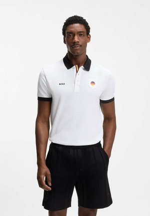 Mężczyzna ubrany w białą koszulkę polo z czarnym kołnierzykiem i emblematem flagi Niemiec, w połączeniu z czarnymi szortami, stojący na jednolitym tle.