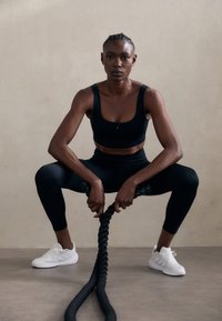 Melna sporta krūšturis un legingi, strukturēta virsma, baltas sporta apavas, turēšana biezas melnas virves, neitrāls fons.