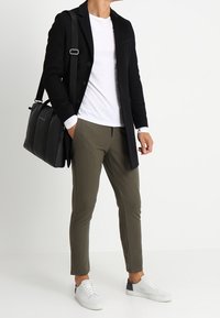 Un homme dans un manteau noir élégant, un t-shirt blanc et un pantalon vert olive tient un sac à dos noir, portant des baskets blanches. Apparence simple et moderne.
