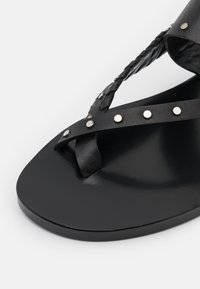 Sandale en cuir noir avec des brides tressées et des clous en argent décoratifs ; semelle plate avec une finition lisse.