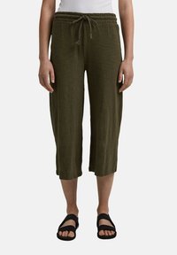 Pantaloni in lino verde oliva, corti, con elastico in vita e cordoncino. Indossati con sandali neri, con punta aperta e design a fascia.
