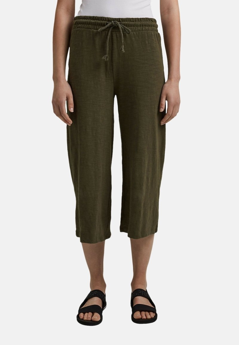 Pantaloni in lino verde oliva, corti, con elastico in vita e cordoncino. Indossati con sandali neri, con punta aperta e design a fascia.